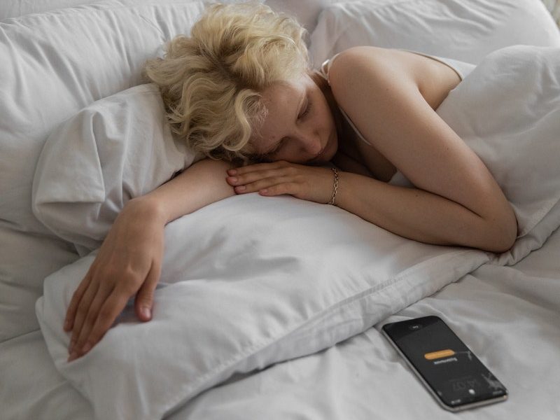 Lazy female resting in bed under soft blanket with alarm on cellphone for waking up